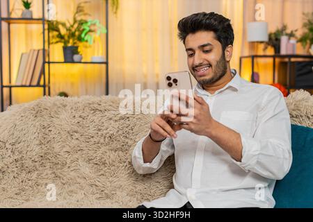 Indian young man at home sitting on sofa focused on smartphone typing text, browsing news, reacting to notifications. Arabian guy using mobile phone for social media, work, texting, digital lifestyle Stock Photo