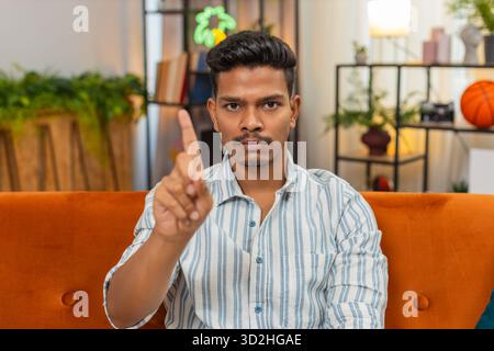 Indian man at home waves finger no no to camera and shows refusal and disagreement with stern mood. Arabian guy on sofa shakes head denies request points finger and shows unhappy sad reject reaction Stock Photo