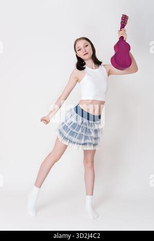 Playful woman ukulele artist and cheerleader holding ukulele, dancing and looking at camera. Cheerful young adult female ukulelist in vest top, short Stock Photo