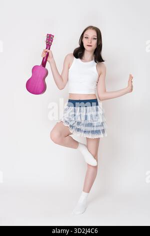 Slender female cheerleader and musician holding ukulele in hand and standing on one leg in full length on white background. 18 year old woman ukulelis Stock Photo