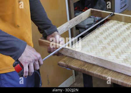 Focused craftsman measuring handmade wooden frame with woven bamboo bottom in workshop. This carpenter uses tape measure for precise DIY furniture pro Stock Photo