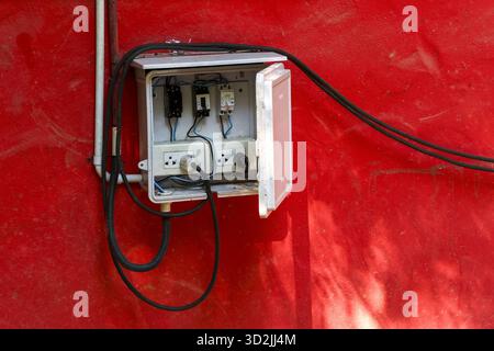 Open electrical box showing messy connection of wire, plug, and socket. This hazardous power supply on red wall has chaotic industrial feeling and uns Stock Photo