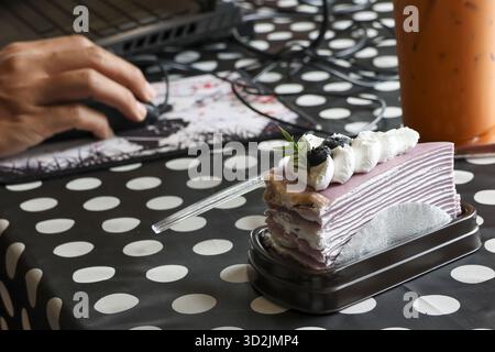 Delicious purple layered cake, sweet dessert, sits on polka dot tablecloth. person hand calmly operates computer mouse, displaying focused work in mod Stock Photo