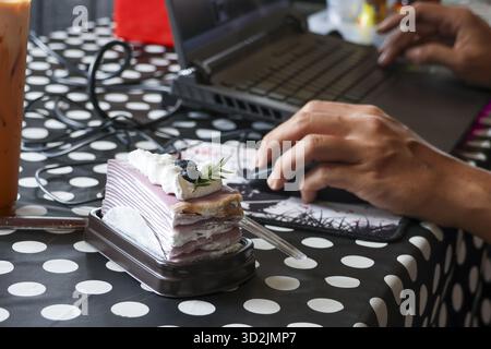 Adult person focused on work, typing and using mouse on laptop. blueberry crepe cake sits nearby, adding sweet moment to busy desk Stock Photo