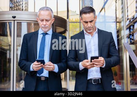 Two businessmen in suits standing outside office, using smartphones seriously. Stock Photo