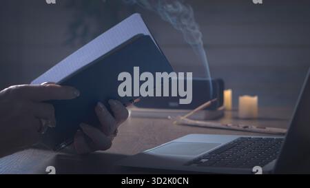 Evening stillness. A pair of thoughtful hands folded near a desktop computer, amber light and incense haze create a mood of gentle reflection Stock Photo