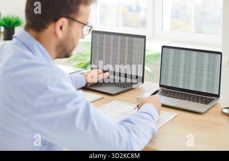 Male focused trader at desk working with two laptop screens checking stock market data using display Stock Photo