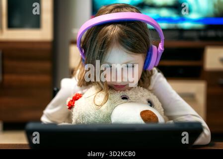 Girl with purple headphones watching tablet closely while holding teddy bear. Image symbolizing childhood technology use, online entertainment and dig Stock Photo
