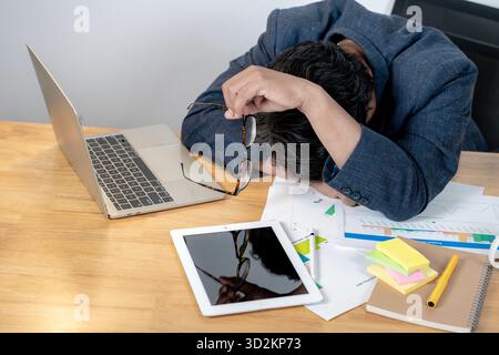 Young Asian businessman sitting at his desk with his hand on his forehead, feeling tired and stressed from heavy workload and long working hours, symb Stock Photo