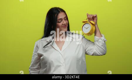 Young woman in white shirt holding yellow clock smiling against isolated yellow background Stock Photo