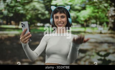 Young hispanic woman with cat ear headphones holds smartphone and beckons hand in forest; friendly invitation. Stock Photo