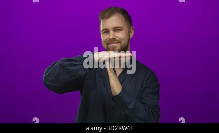 Man young caucasian with beard and hands making timeout gesture while smiling in purple studio backdrop; playful pause. Stock Photo