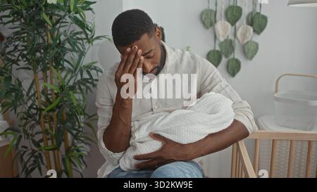 Man holding swaddled baby, hand to forehead in building nursery by crib and plant; fatigue tenderness bonding. Stock Photo