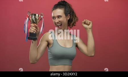 Woman holds trophy with fist raised in studio against a vivid red backdrop, smiling broadly; pride celebration. Stock Photo