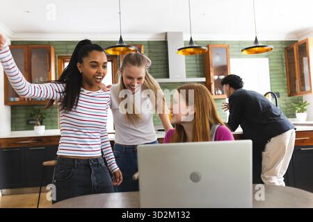 Teenagers celebrating success while using laptop in modern kitchen together. Technology, celebration, friendship, happiness, teamwork, lifestyle || Model released Stock Photo