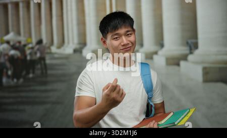 Young chinese man gestures on a city street with columns in the background, holding colorful notebooks for studying. Stock Photo