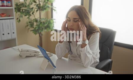 Woman doctor using tablet in hospital workplace surrounded by office equipment and natural light, focused on client consultation. Stock Photo
