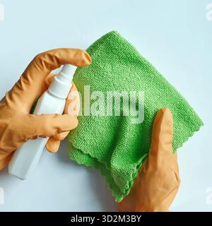Hands wearing orange gloves cleaning with spray bottle and green microfiber cloth on light blue background. Stock Photo