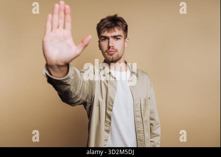 Young man with outstretched hand signaling stop against beige background. Stock Photo