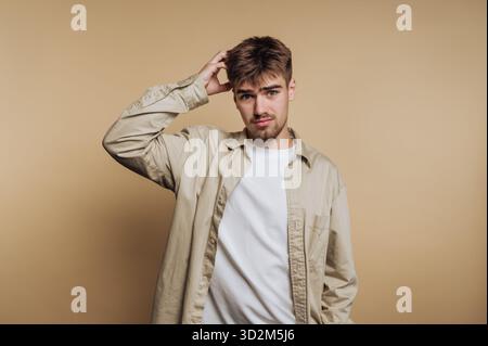 Young man in casual clothing scratching his head, beige background. Stock Photo