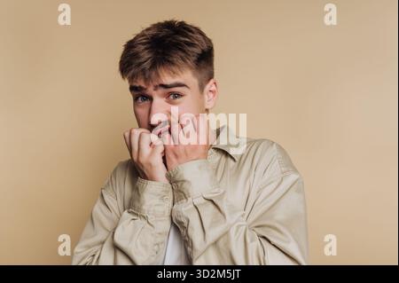 Young man biting nails, looking anxious against beige background. Stock Photo