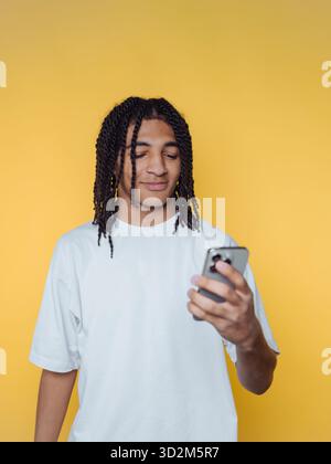 Young man with braided hair using smartphone against yellow background. Stock Photo