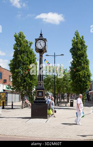 A classic clock tower stands in a lively town square under a clear blue sky, with festive bunting and pedestrians enjoying a bright summer day near sh Stock Photo