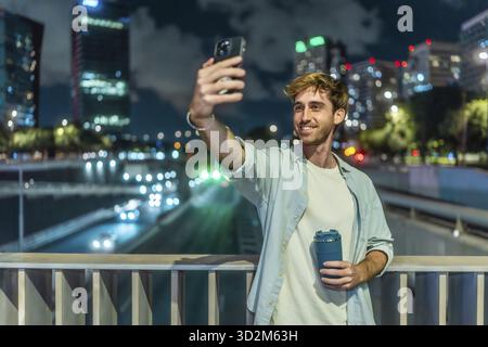 Young man smiling and taking a selfie with a smartphone, holding a reusable coffee cup on an elevated city bridge at night, with traffic lights blurri Stock Photo