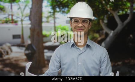Young hispanic man wearing hardhat reviews blueprints while smiling near building; planning confidence. Stock Photo
