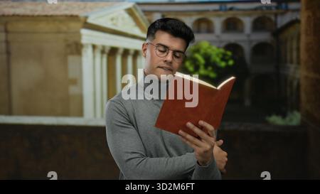 Young man reading a book in front of an old university building, wearing glasses and a turtleneck, focused on studying in an academic setting. Stock Photo
