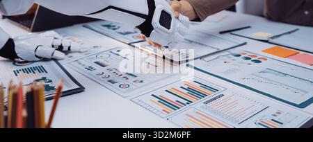 A robot shakes hands with a person in a modern office while surrounded by data charts and documents, symbolizing the future of technology and human co Stock Photo