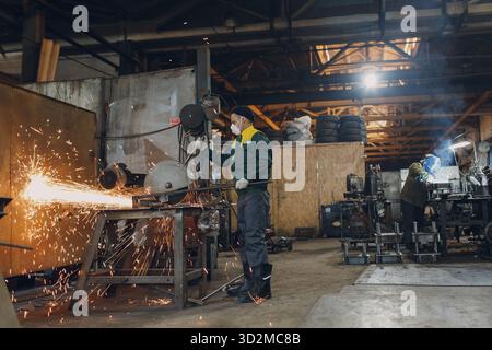 Workers working with metal construction on plant. Metal processing with big angle grinder disk saw and welder welding. Sparks in metalworking at facto Stock Photo