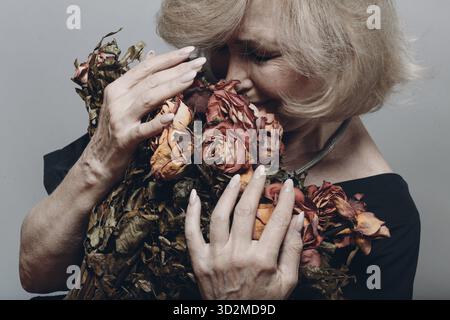 Upset elderly woman crying and holding withered dry old rose flowers bouquet Stock Photo
