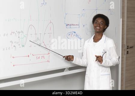 African American smiling woman math teacher stands at black board with pointer Stock Photo