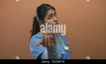 Young hispanic woman doctor with stethoscope beckons with outstretched hand in studio; invitation command. Stock Photo