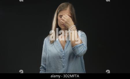 Young blonde woman pinches bridge of nose and closes eyes under harsh lighting in black studio; disgust. Stock Photo