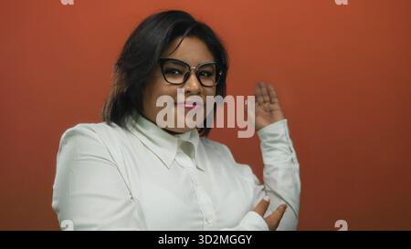 Young woman in glasses with hands back poses confidently against a vibrant red background showcasing her stylish attire and positive demeanor. Stock Photo