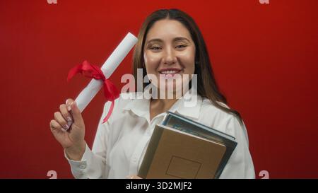 Young hispanic woman holds diploma with red ribbon and stack of books in studio; proud achievement. Stock Photo
