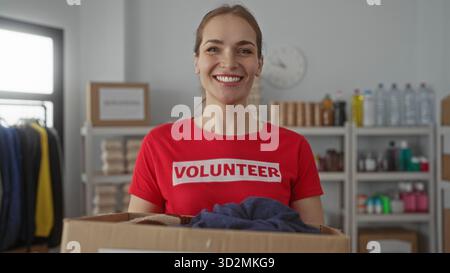 Woman holds large cardboard donations box with hands in building filled with shelves and clothing donations; generosity. Stock Photo