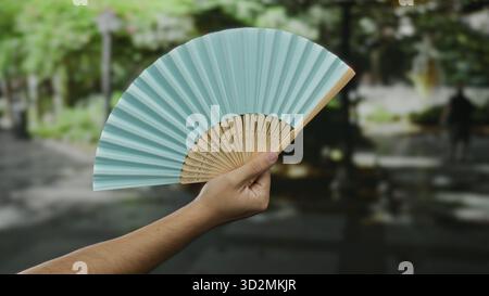 Man holding blue fan outdoors in park setting on sunny day showcasing traditional accessory with blurred green background. Stock Photo