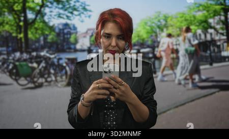Young hispanic woman using smartphone with fingers tapping screen on amsterdam street beside canal and parked bikes; concentration. Stock Photo
