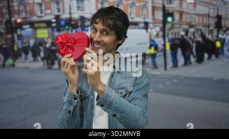 Man holding red heart shaped gift box near smiling face in bustling crowded urban street; love romance. Stock Photo