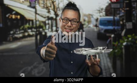 Young chinese man holding drone and giving thumbs up on street, smiling and looking at camera; confidence innovation. Stock Photo