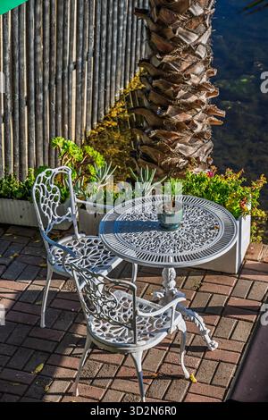 Table and two chairs made of white, curved metal. Sitting area on a wooden terrace on the river bank. Stock Photo