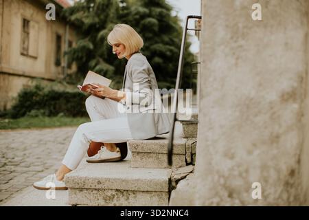 A woman reads a book while sitting on stone steps outside an old building. She wears a light blazer and white pants, surrounded by greenery in a peace Stock Photo