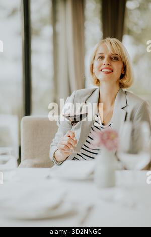 A young woman sits at a beautifully set table in a well-lit restaurant, smiling as she holds a glass of red wine. The atmosphere is relaxed and inviti Stock Photo