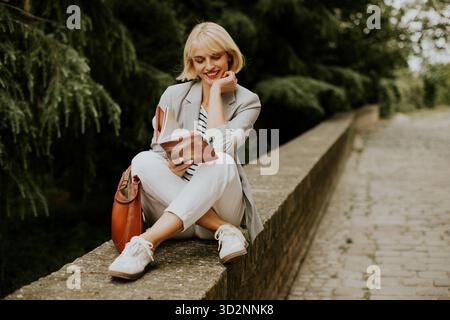 A young woman with blonde hair sits outside and reads a book. She is sitting on a ledge with trees in the background and smiles as she is reading. Stock Photo
