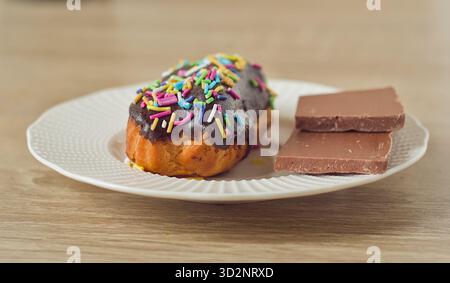 A delicious chocolate dessert on a white plate. an eclair cookie is covered in chocolate and sprinkles, next to squares of milk chocolate bar Stock Photo