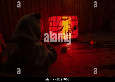 Child in warm clothing looking at glowing red illuminated cube with Halloween spider and bat decorations on wooden terrace, soft night light creating Stock Photo