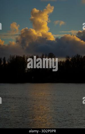 Dramatic vertical clouds above Daugava River and trees in Riga during golden evening hour showing autumn reflection, calm horizon, and soft atmospheri Stock Photo